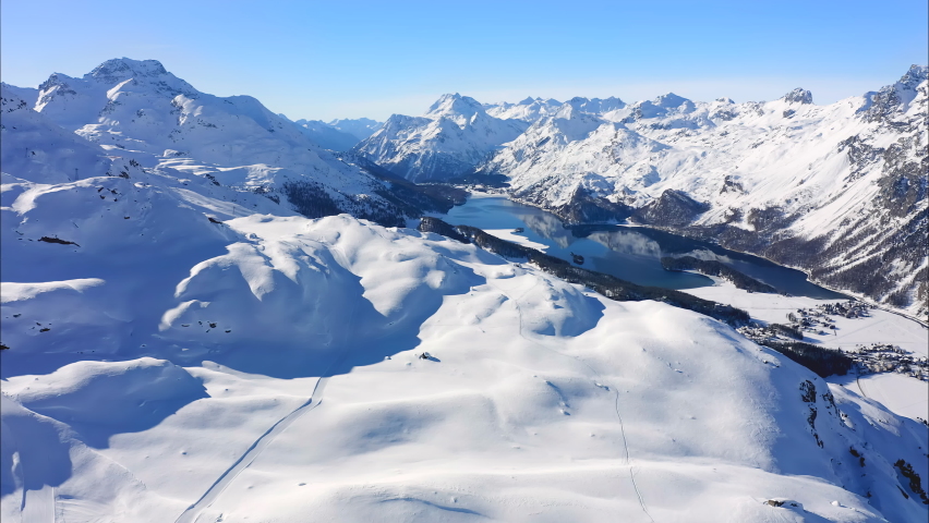 St. Moritz, Switzerland: Aerial view of famous alpine ski resort Saint Moritz in winter Swiss Alps, snow on mountain slopes, Lake Sils on sunny day with clear blue sky - landscape panorama of Europe Royalty-Free Stock Footage #1081888385