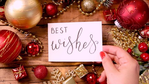 Woman's hand put a Christmas card with the handwritten text BEST WISHES on a table with Christmas decorations around top view