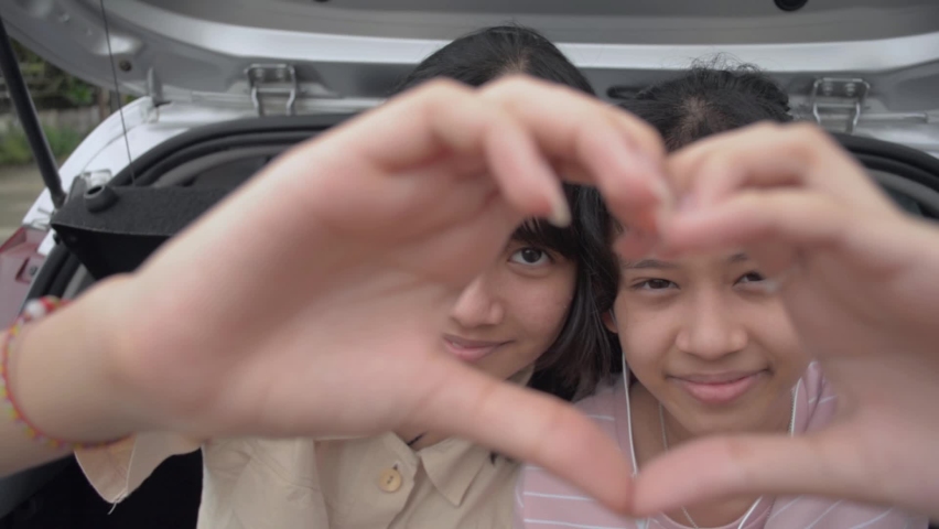 Happy two Asian teen sisters making hand gesture heart shape and smiling while looking at camera. Teen girls sitting in the rear side of hatchback car. Bonding relationship of sibling family.