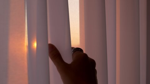 Light white matte tulle sways in the wind. A woman's hand with a ring on her finger reaches out to the curtain behind which the rays of the setting sun shine through.