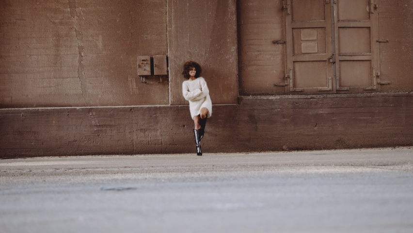 Wide Slow Motion Shot Towards Smiling Woman With Windswept Afro In Woollen Dress And High Heeled Boots Leaning Against Wall In Street Royalty-Free Stock Footage #1081909370