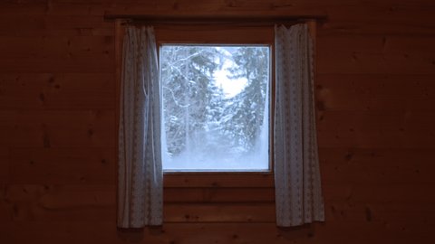 Snowfall and snow-covered trees seen through a small window with curtains in a wooden wall of a mountain hut in the alps.
