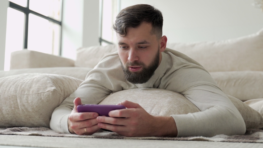 Portrait of an excited young bearded man lying on the floor and playing games on a mobile phone