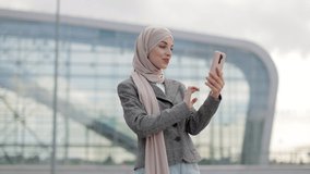 Likable pretty young Muslim woman greeting her friend with waving hand, having video call outside. Arab businesslady smiling and talking with friend near modern building