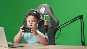 Camera And Asian Little Boy Playing Video Game With Mobile Phone Then Celebrating While Live Stream On Green Screen 
