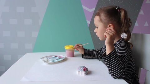 A little curly girl of European appearance of 3-4 years old sits at the desk and paints a plaster figure of a hippo. Education concept. Home hobby and fun game.