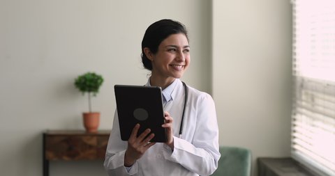 Smiling young beautiful female indian ethnicity doctor general practitioner using digital computer tablet, managing appointments or healthcare treatments, giving distant professional medical help.