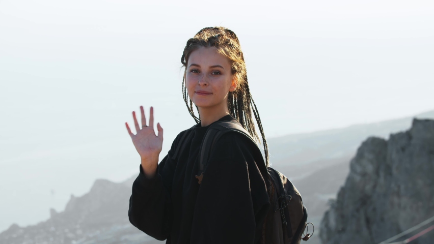 Young pretty woman hiker with dreadlocks standing on a background of mountains and waving her hand to a camera