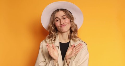 Pretentious blonde curly hair woman in trench coat and hat clapping, and looking annoyed over yellow background. Girl applauds with a grin, sarcasm sarcastically clapping her hands.