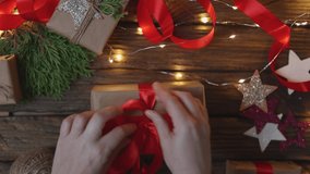 female woman hands wrapping Christmas presents gift box with brown craft paper, tying red ribbon bow on wooden desk with new year ornaments: stars light garland and green coniferous. top view DIY home - Powered by Shutterstock - Get 15% off with code: PIKWIZARD15