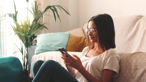 Serious woman in eyeglasses text messaging using mobile phone while sitting on floor in the living room of her house. Beautiful lady spending leisure time using smartphone at home
