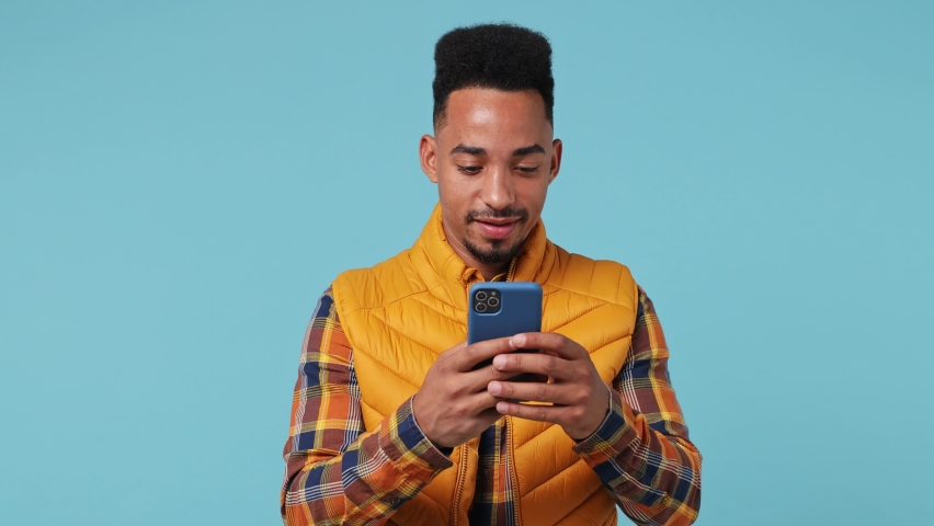 Surprised young african american†man 20s wears yellow shirt waistcoat hold use point on mobile cell phone say wow yes found out big win isolated on plain pastel light blue background studio portrait