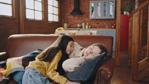 Young mother in casual clothes fell asleep with book on couch in kitchen of wooden country house. Little daughter coming up to her, covering mother with plaid and hugging. Happy family, relationship