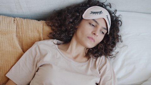 Portrait of pretty woman putting on beautiful sleep mask yawning and sleeping in comfortable bed at home. Lifestyle and relaxation concept.