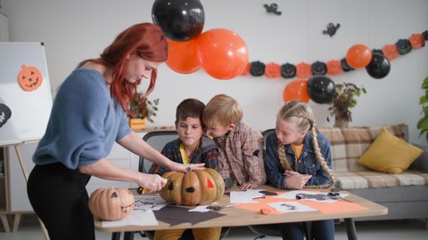 home family halloween party, young mom carves eyes on a pumpkin, curious children sitting at table watching mom on background of garlands and balloons