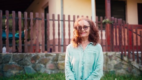 Handheld cinematic shot. Portrait of a mature adult woman in glasses standing against the background of her country house on a windy sunny day and looking at the camera.
