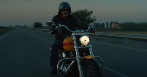 Cinematic shot of active mature retired senior motorcyclist with protective helmet and leather jacket is riding his custom motorbike in high speed along country side road at dusk. 