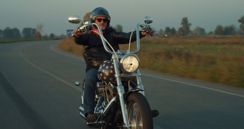 Cinematic shot of active mature retired senior motorcyclist with protective helmet and leather jacket is riding his custom motorbike in high speed along country side road at sunset. 