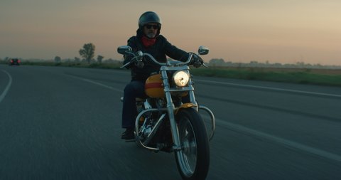 Cinematic shot of active mature retired senior motorcyclist with protective helmet and leather jacket is riding his custom motorbike in high speed along country side road at dusk. 