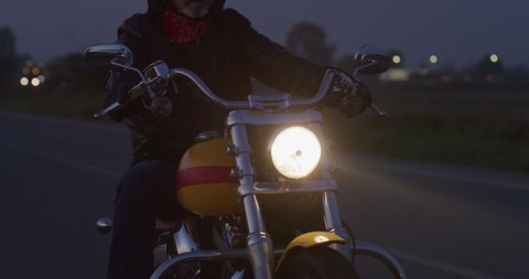 Cinematic close up shot of active mature retired senior motorcyclist with protective helmet and leather jacket is riding his custom motorbike in high speed along country side road at dusk. 