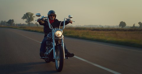 Cinematic shot of active mature retired senior motorcyclist with protective helmet and leather jacket is riding his custom motorbike in high speed along country side road at sunset. 