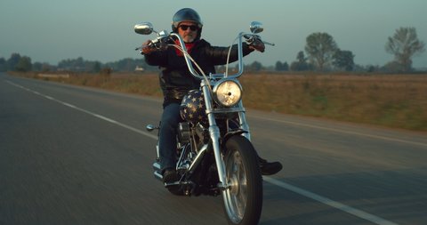 Cinematic shot of active mature retired senior custom motorcyclist with protective helmet and leather jacket is riding his motorbike in high speed along country side road at sunset. 