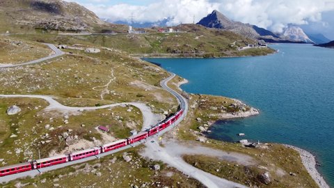 Aerial drone footage of a train travelling along the lake at the summit of the Bernina pass in the alps in Canton Graubunden in Switzerland. Shot with a following motion. 
