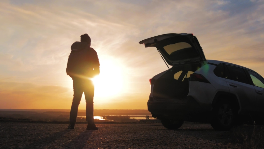 Back view of a silhouette man with backpack arms wide open to the sides at sunset. Young male traveler is enjoying beautiful landscape during road trip on suv. Road trip, travel and people concept.