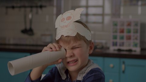 Kid playing pirate's game with paper telescope and headwear with skull and crossbones. Preschool boy searching for sweets with pirate's telescope. Child eating cookies.