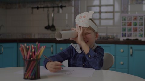 Kid playing pirate's game with paper telescope and headwear with skull and crossbones. Preschool boy drawing with color pencils sitting alone at home.