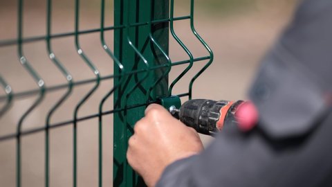A worker screws a green mesh to a metal fence with a screwdriver. Installation of a fence in close-up.