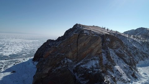Aerial orbital shot of cape Khoboy, Olkhon island. Tall rocks in frozen lake Baikal with many people and cars around. Popular touristic destination. Winter landscape. Panoramic view