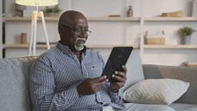 Distant communication. Positive african american grandfather video chatting with family, waving hand to digital tablet, sitting on sofa at home. slow motion, empty space