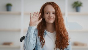 Disability and positive. Cute little impaired redhead girl sitting in wheelchair and waving hand to camera, saying hello to interlocutors, webcam pov portrait, slow motion