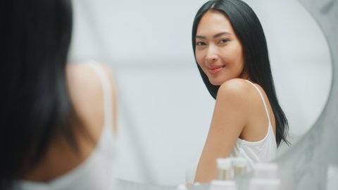 Beautiful Asian Woman Touches Her Perfect Soft Shoulder, Neck, Sensually Smiles in the Mirror. Happy Female Enjoying Her Beauty Routine. Wellness Natural Cosmetic Skincare Products. Focusing Portrait