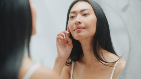 Beautiful Asian Woman Her Perfect Soft Face Skin, Uses Advanced Organic Cream, Smiles in the Mirror. Happy Female Enjoying Her Morning Beauty Routine. Wellbeing, Natural Beauty, Cosmetic Products