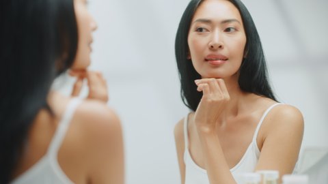 Beautiful Asian Woman Touches Her Perfect Soft Face Skin and Lush Black Hair, Smiles in the Mirror. Happy Self Loving Female Enjoying Her Wellbeing, Natural Beauty, Organic Skincare Cosmetic Products