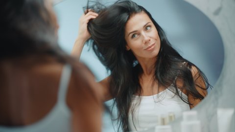 Beautiful Early Middle Aged Woman Looks into Bathroom Mirror Touches Her Lush Black Hair, Admires Her Looks. Concept for Happiness, Wellbeing, Natural Beauty, Organic Skin Care Products