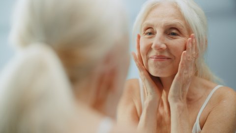 Beautiful Senior Woman Looks into Bathroom Mirror Touches Her Neck, Happily Smiles, Enjoy Her Looks and Morning Routine. Dignity and Grace in Old Age. Wellbeing, Skin care, Beauty Products