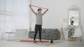 Handsome Young Man in a Gray T-Shirt and Black Shorts Does Sports at Home Online. Active fitness male showing exercise for back and arms muscle using rubber band