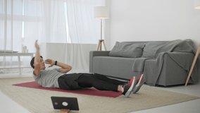 Handsome Young Man in a Gray T-Shirt and Black Shorts Does Sports at Home Online. Sportive Male training abdominal muscles on floor indoor. Cross crunches.