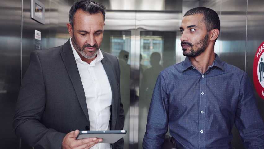 Two business colleague having a conversation while walking. Businessman and boss discussing together with digital tablet in a modern office. Business men discussing work coming out of an elevator. Royalty-Free Stock Footage #1081987025