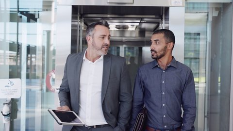 Two business colleague having a conversation while walking. Businessman and boss discussing together with digital tablet in a modern office. Business men discussing work coming out of an elevator.