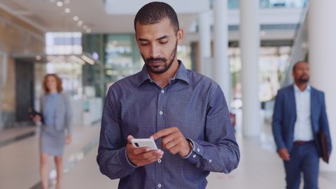 Happy young businessman using smartphone and looking at camera. Successful mixed race business man smiling while reading message on cellphone. Middle eastern man using mobile phone in modern workplace