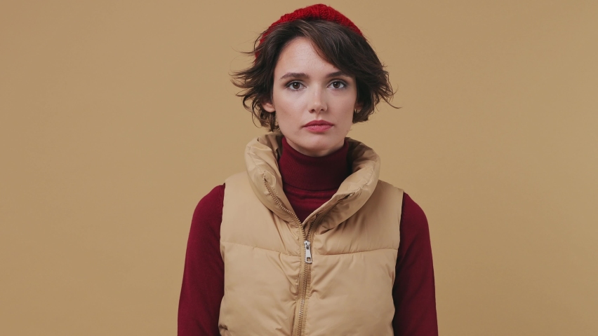 Cold young brunette woman 20s wears red hat shirt waistcoat rub hands in mittens try to warm up blow look around isolated on plain pastel beige background studio portrait. Autumn spring season concept