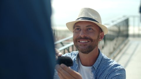 Front view of smiling Caucasian gay proposing to lover outdoors. Slow motion of happy and handsome homosexual man wearing hat holding box with engagement ring in front of soulmate. LGBT, love concept