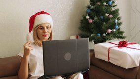 Female in Santa Claus hat dancing with her hands sitting on sofa in decorated living room. Christmas holidays concept. Female person chatting with friends, family online