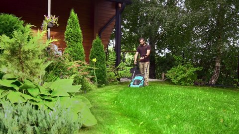 Lawn care. Mowing grass with an electric lawn mower. A young man mows the grass with a lawn mower with a grass collector on a sunny summer day. Beautiful landscape design in the garden.