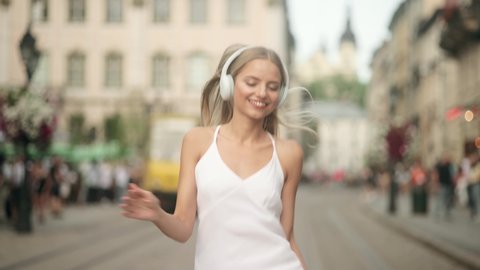 Slow motion of happy young blonde woman in wireless headphones dancing singing outdoors in city street having fun alone. Young adult happy woman in a white dress dancing in downtown very emotional