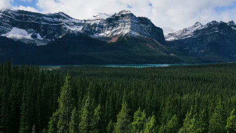 Drone Shot of Scenic Mountains with snow peaks and pine woods in Banff National Park on a sunny day. Lake with turquoise water. Aerial video in mountain over a forest in the Rocky Mountains in Canada.